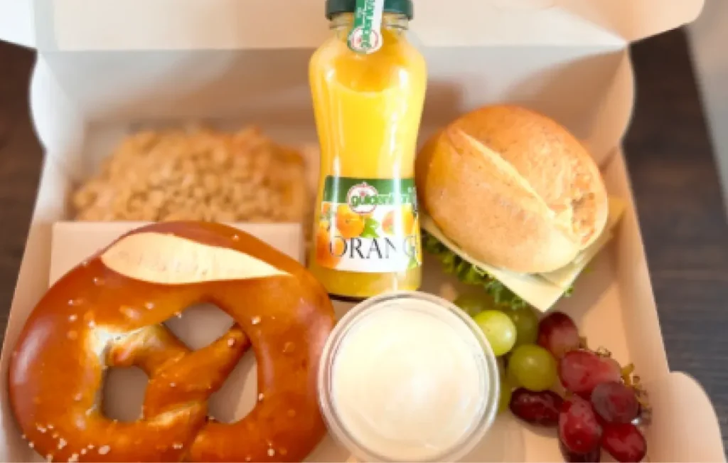 Geöffnete Frühstücksbox mit Brezel, belegtem Brötchen, Streuselteilchen, Joghurt, Trauben und Orangensaft.