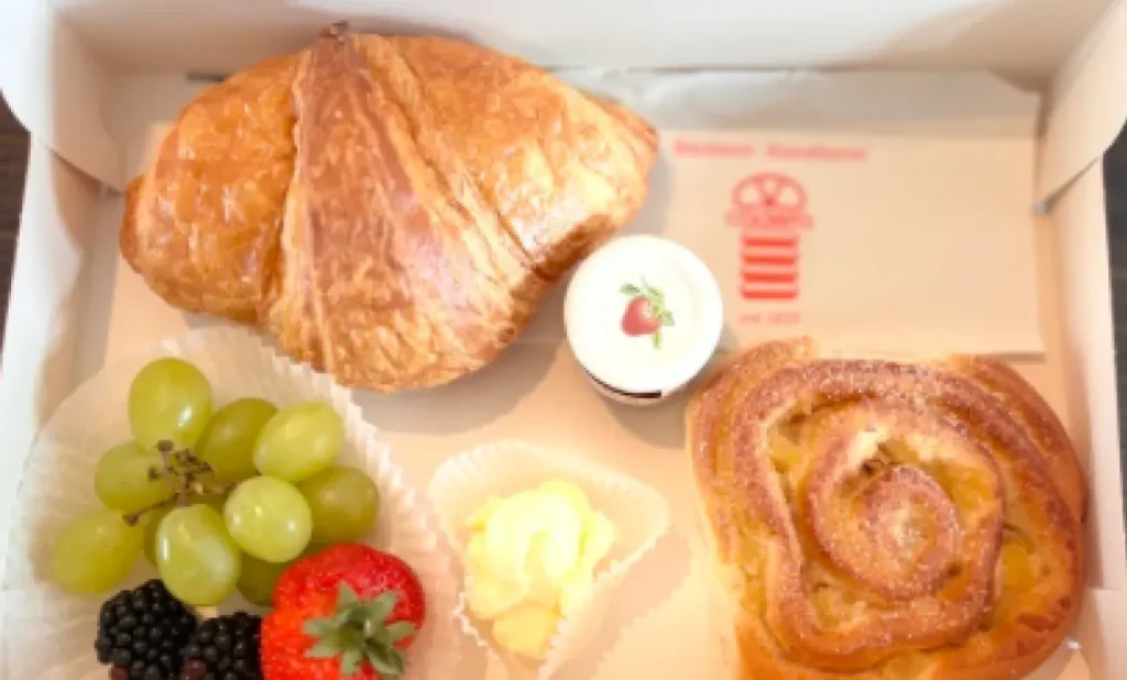 Geöffnete Frühstücksbox mit Croissant, Apfelteilchen, Trauben, Beeren, Butter und Marmelade.