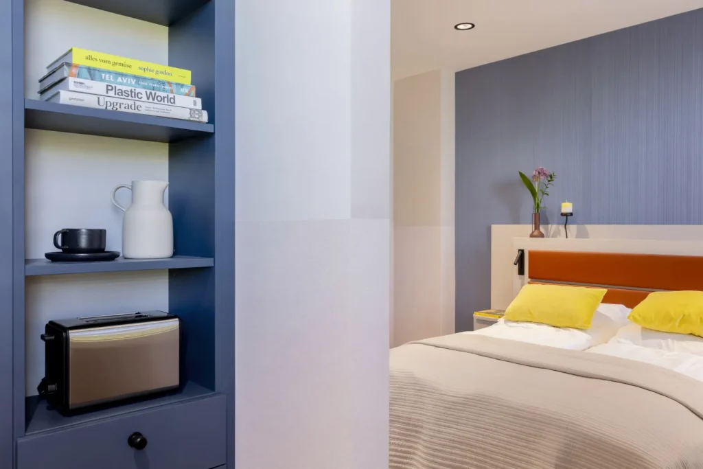 Example image: Blue shelf with books, coffee cup and toaster; behind the bed is a placeholder for ipartment Fürth