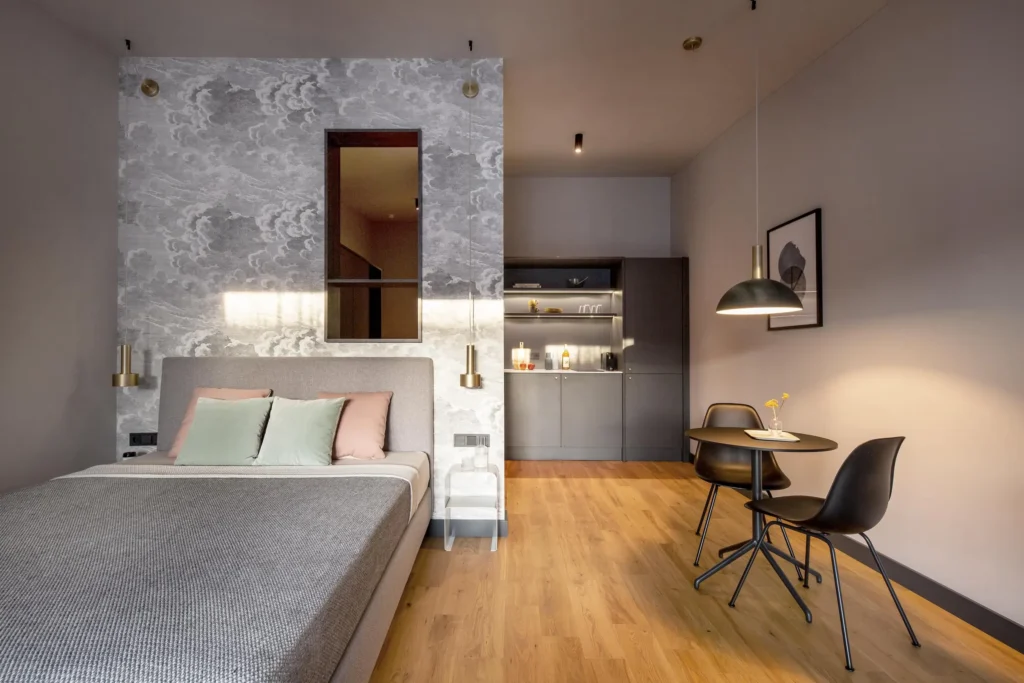 Double bed in front of a cloud wallpaper, kitchenette and round dining table in the Medium apartment, ipartment Hamburg Eimsbüttel