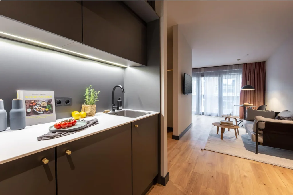 Kitchenette in the hallway with a view of the living area with a sofa and a window in the Medium apartment, ipartment Hamburg Eimsbüttel