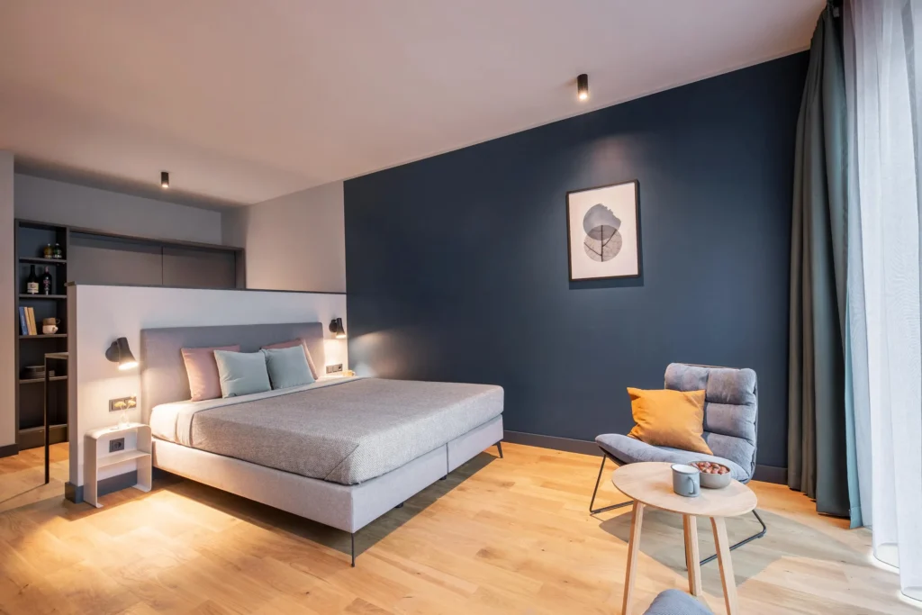 Sleeping area with box spring bed, armchair and floor-to-ceiling window in a blue-painted room, ipartment Hamburg Eimsbüttel
