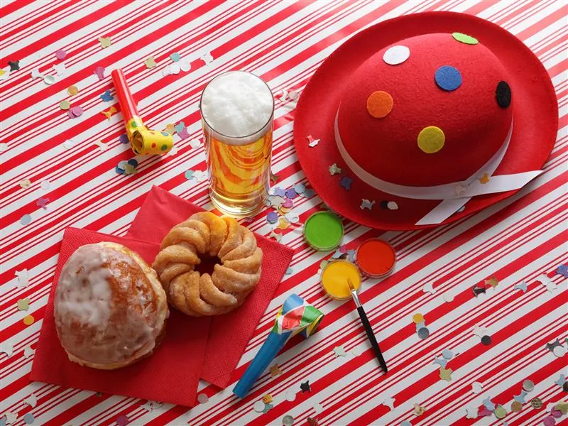 Narrenkappe mit bunten Punkten, Kölschglas, Berliner, Schmalzgebäck, Partytröten und Schminkfarben auf rot-weiß gestreifter Unterlage mit Konfetti