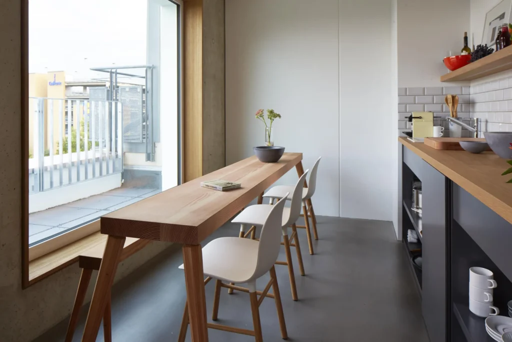 Long wooden table with white chairs at the floor-level window with balcony in the XL apartment, ipartment Cologne Mülheim
