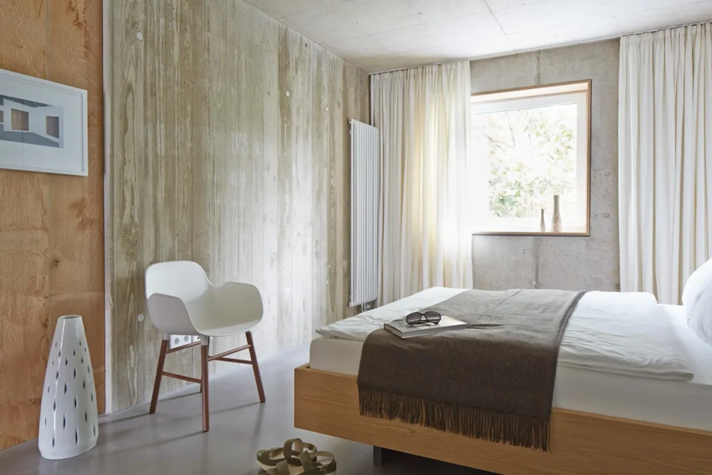 Sleeping area with double bed, see-through concrete wall and window with curtain in the XS apartment, ipartment Cologne Mülheim