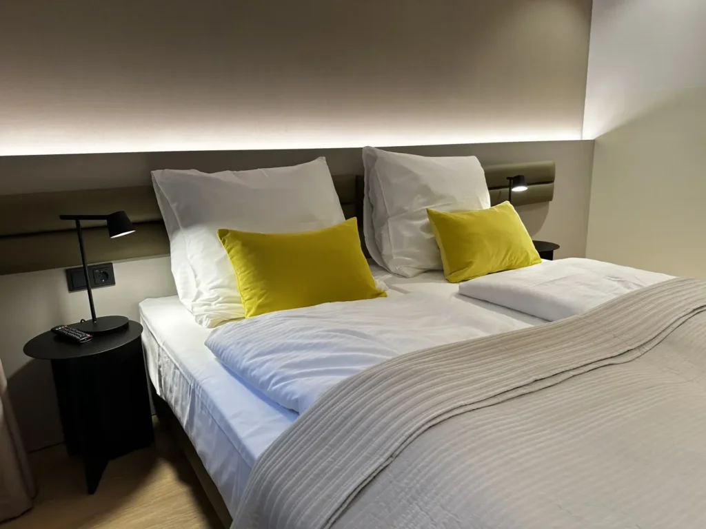 Doppelbett mit weißer Bettwäsche und gelben Kissen, darüber eine indirekt beleuchtete Wandblende