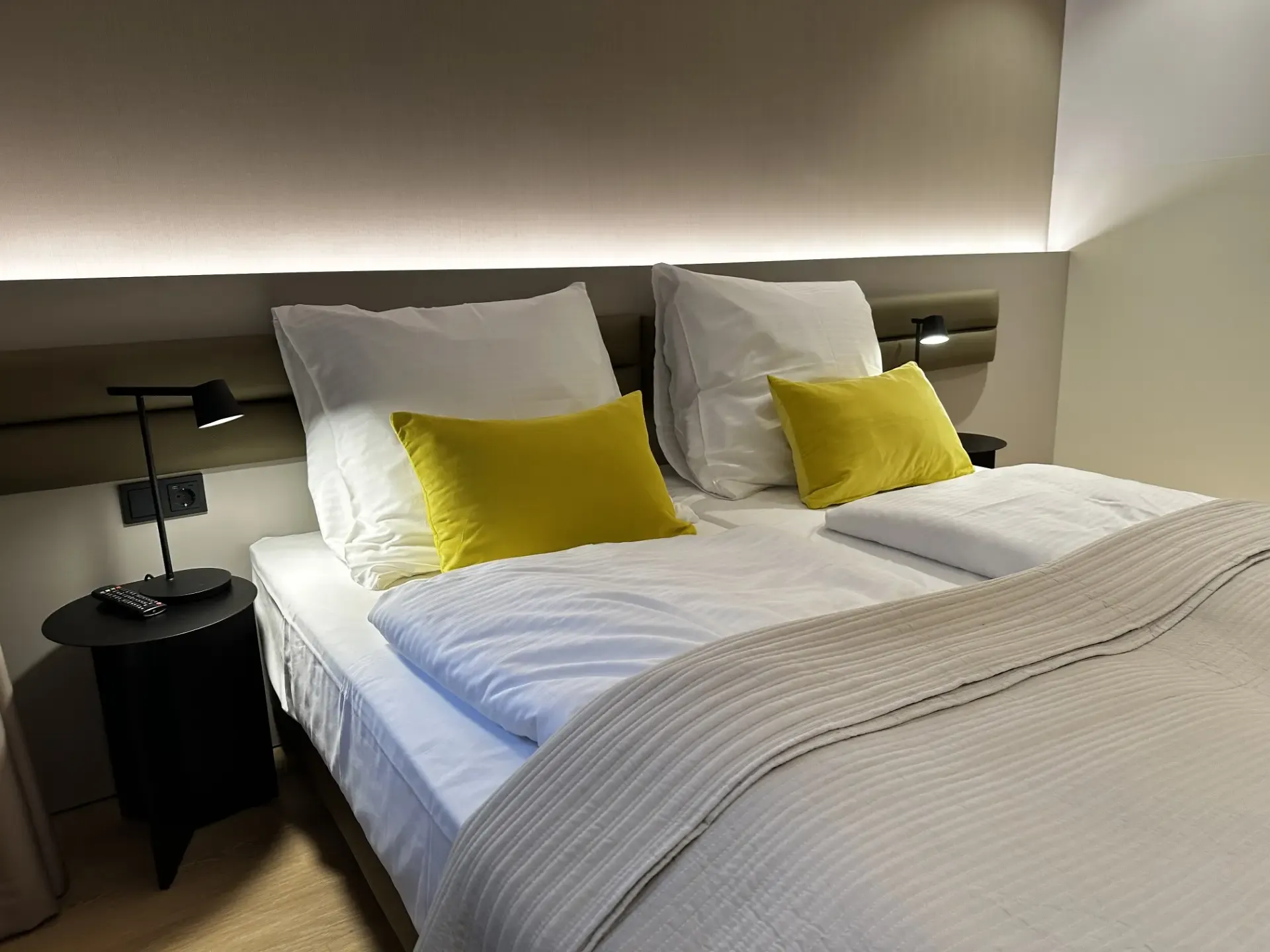 Doppelbett mit weißer Bettwäsche und gelben Kissen, darüber eine indirekt beleuchtete Wandblende