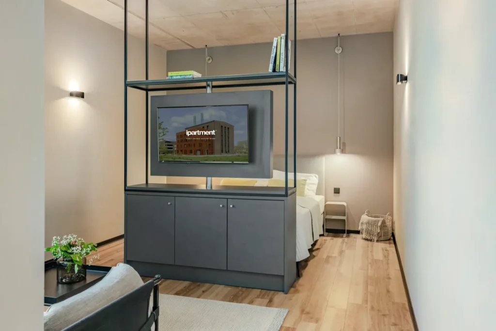 Sleeping area with bed, wall lights and TV on a shelf element in the Medium apartment, ipartment Leverkusen