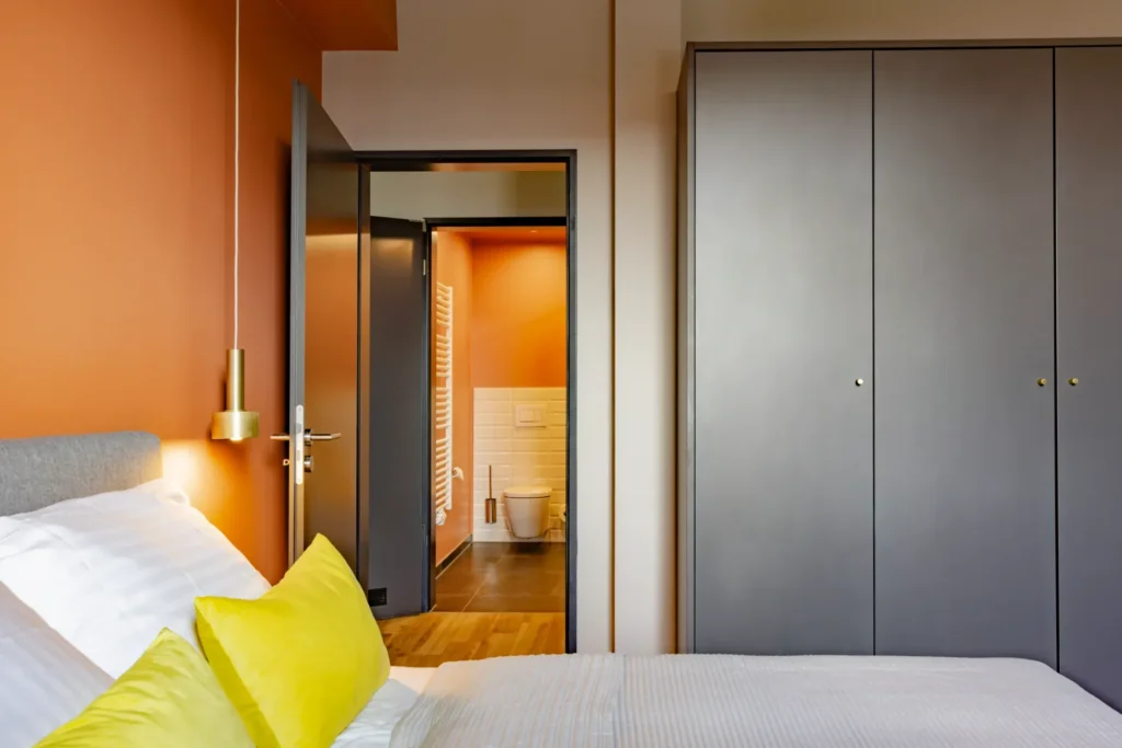 Bed with yellow pillows in front of an orange wall, wardrobe and open door to the bathroom in the Xtra Luxury apartment, ipartment Leverkusen