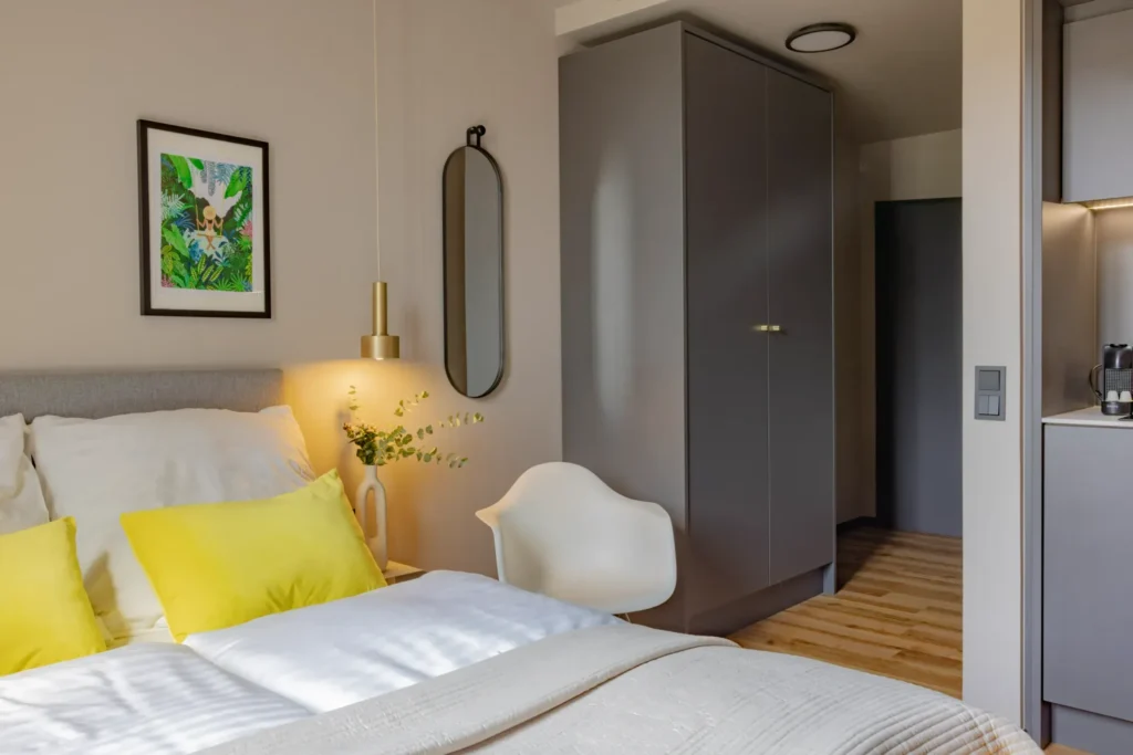 Bett mit gelben Kissen, Kleiderschrank und Blick zur Küchenzeile im Apartment Xtra Smart, ipartment Leverkusen