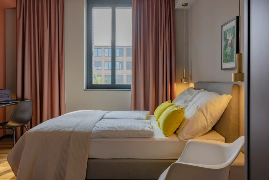 Sleeping area with bed, pink curtains and windows; in the background is the work area in the Xtra Smart apartment, ipartment Leverkusen