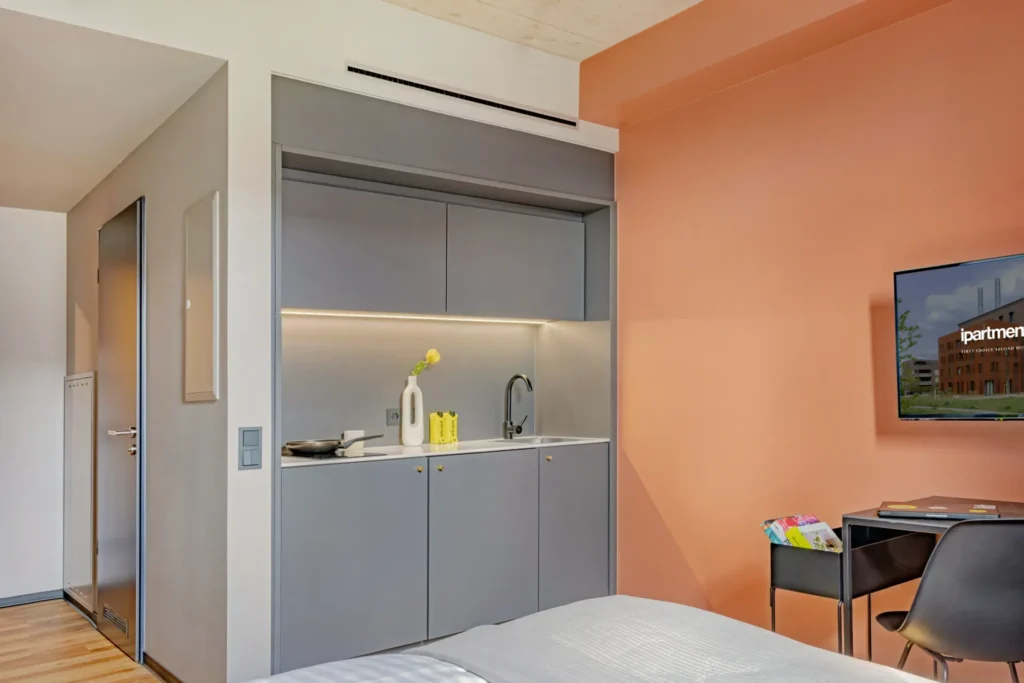 Compact kitchenette with sink, orange wall, TV and desk in the Xtra Smart apartment, ipartment Leverkusen