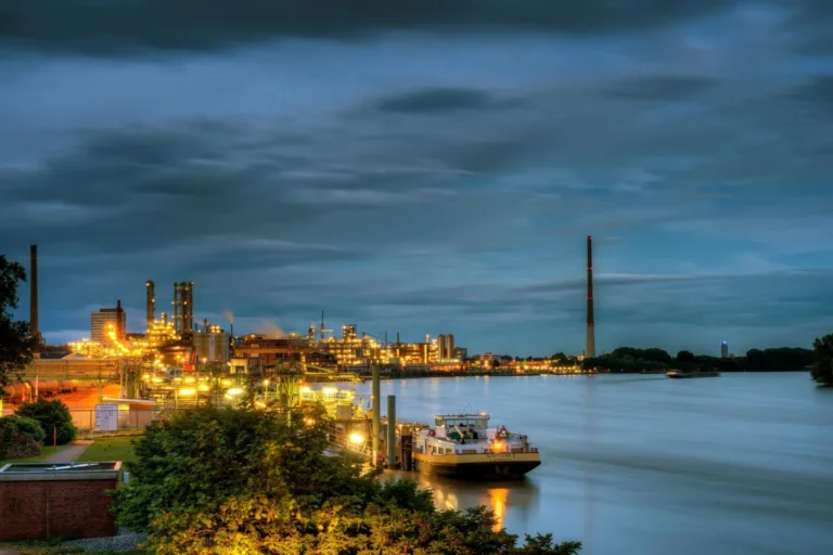 Illuminated industrial plant with chimneys and a tugboat on the river at dusk, Leverkusen
