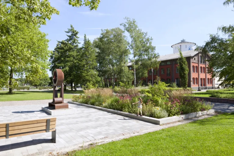 Green area with a flowerbed, sculpture and bench in front of a red brick building in Leverkusen
