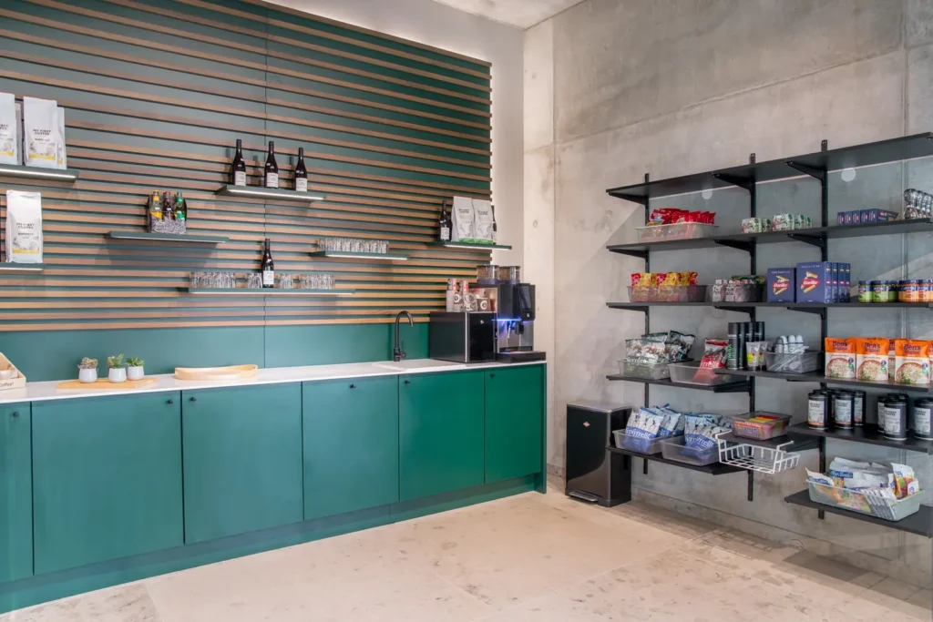 Lounge mit grüner Küchenzeile und Kaffeemaschine, daneben schwarze Regale mit Snacks und Basics vor einer Betonwand.