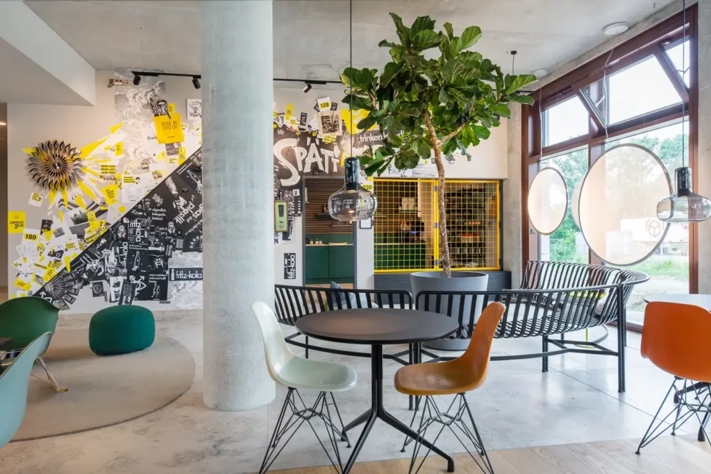 Lobby mit Sitzecken, einer großen Zimmerpflanze und einer Wand mit gelb-schwarzer Collage, im Hintergrund ein Regal mit Snacks und Getränken.