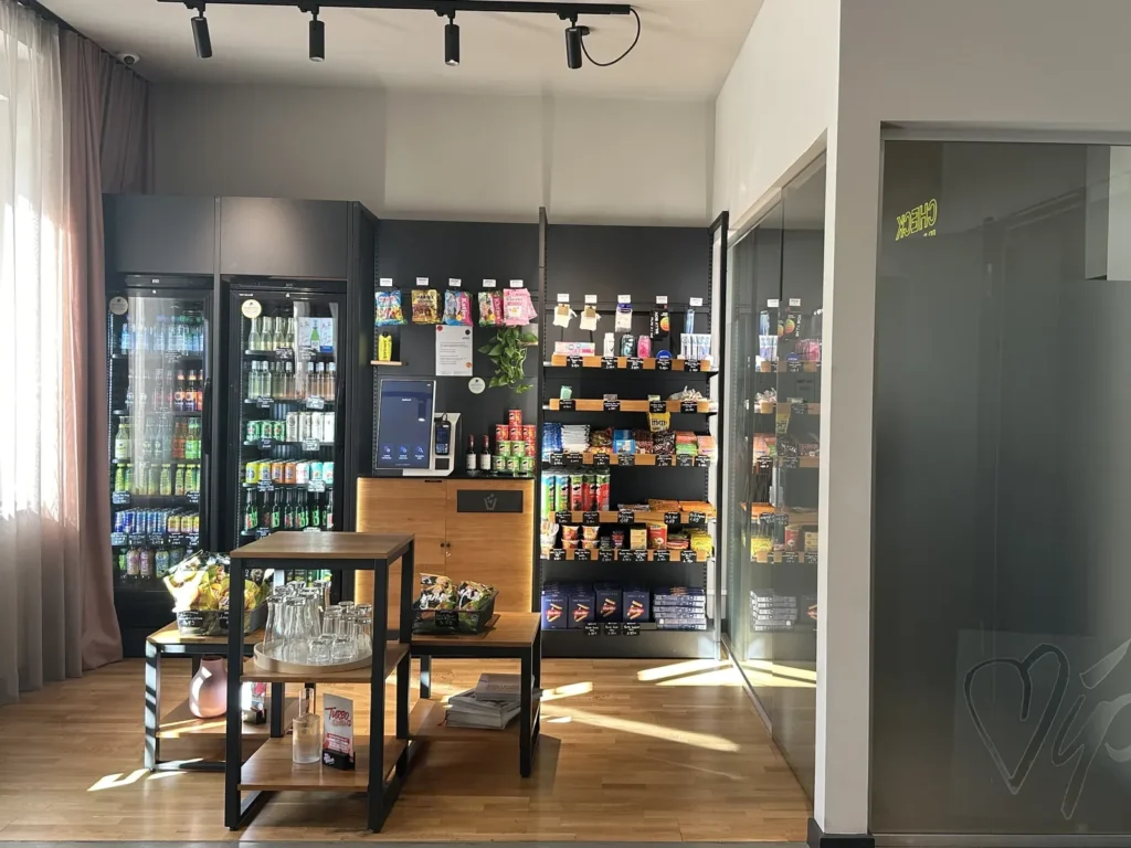 Self-Service-Shop in der Lounge mit Kühlschränken, Regalen voller Snacks und einem Bezahlterminal, davor kleine Holztische mit Gläsern.