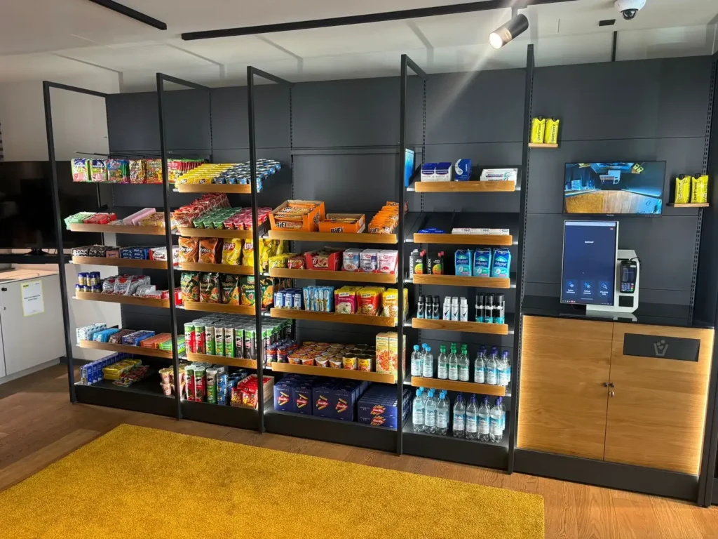 Regale mit Snacks und Wasserflaschen an einer dunklen Wand mit Bezahlterminal, davor ein gelber Teppich.