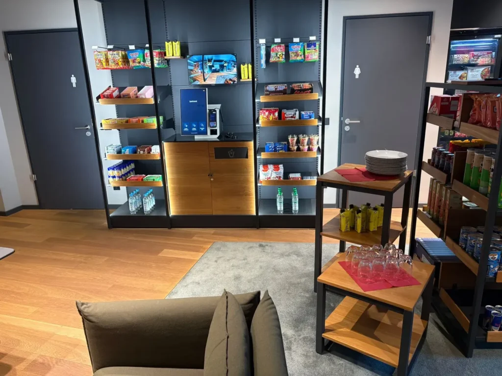 Regale mit Snacks und Getränken an einer dunklen Wand mit Bezahlterminal, davor Tische mit Gläsern und ein Sofa.