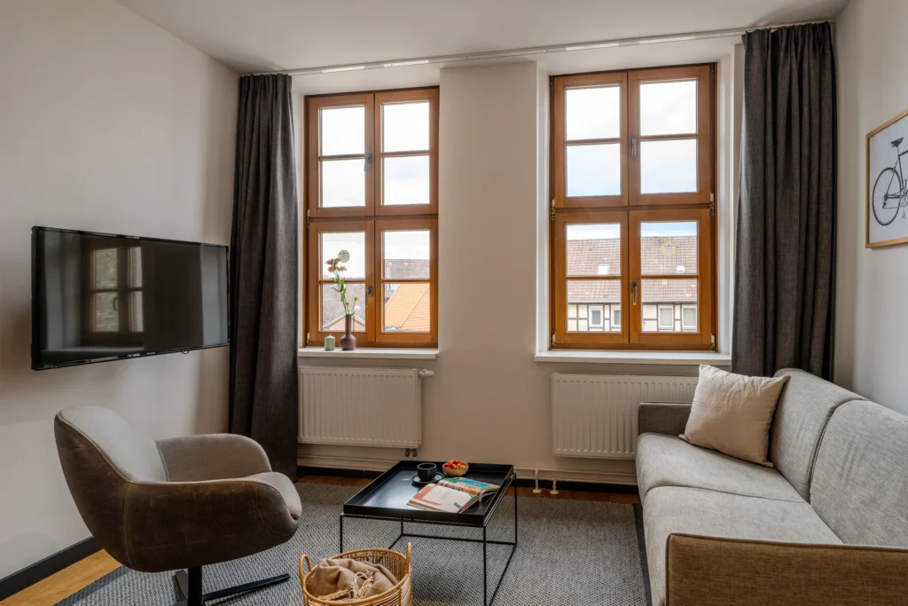 Living room with sofa, armchair and two wooden-frame windows with balcony, ipartment Wolfsburg