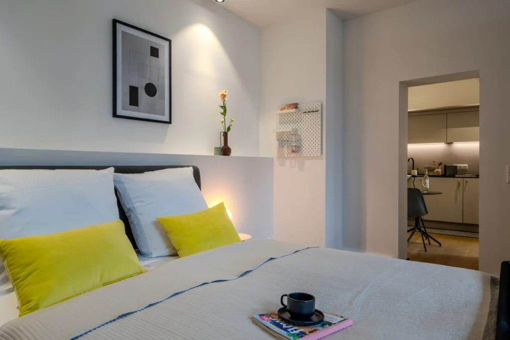 Double bed with yellow pillows, view through the door to the kitchen area, ipartment Wolfsburg