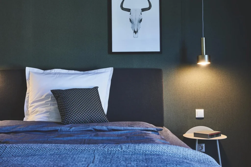 Bed with blue bedding and pendant light in front of a dark wall in the XL apartment, ipartment Wolfsburg