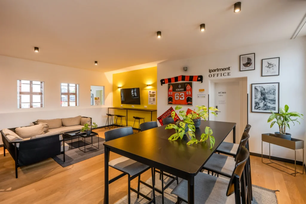 Lounge with long table, sofa, TV and yellow wall next to the office, ipartment Wolfsburg