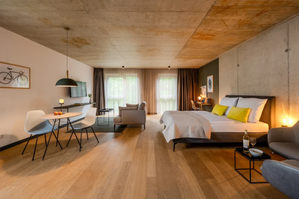 Großer Raum mit Sichtbetondecke: Doppelbett, Sofa, Esstisch und Schreibtisch, ipartment Wolfsburg