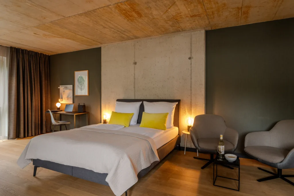 Double bed in front of the view-proof concrete wall, two armchairs and a desk next to it, ipartment Wolfsburg
