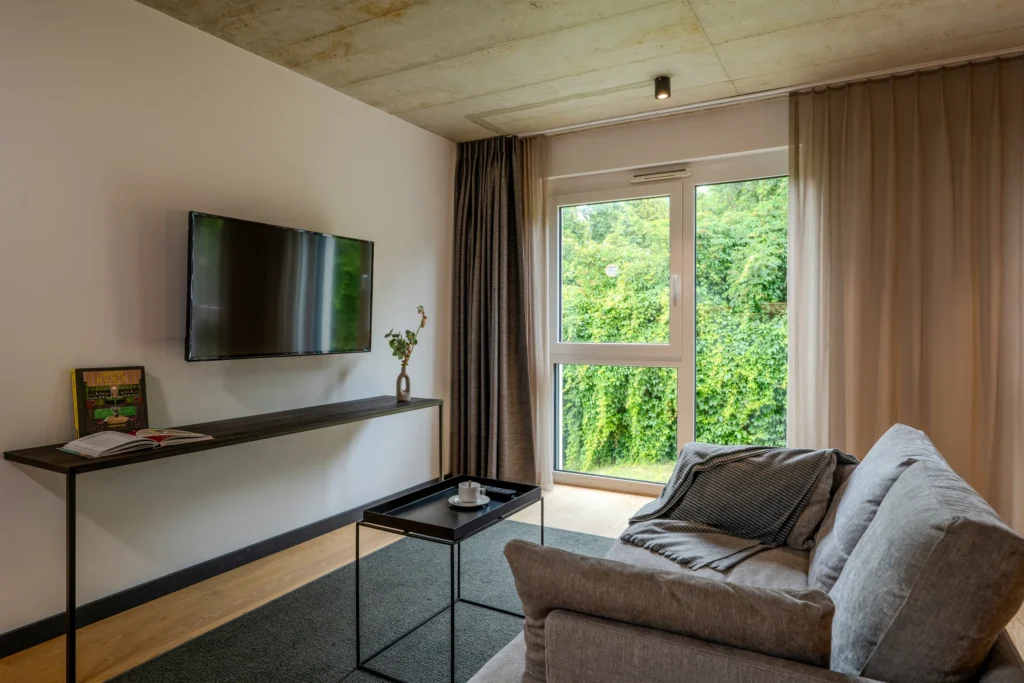Living area with sofa, TV and floor-to-ceiling window to the greenery, ipartment Wolfsburg