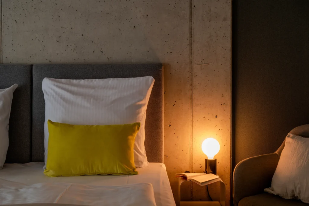 Head of the bed with a yellow cushion in front of visible concrete, next to it a bright nightstand lamp, ipartment Wolfsburg