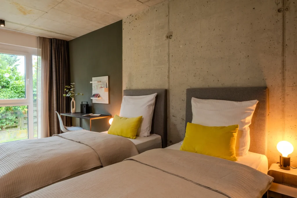 Two single beds in front of a view-proof concrete wall, behind that a desk by the window, ipartment Wolfsburg