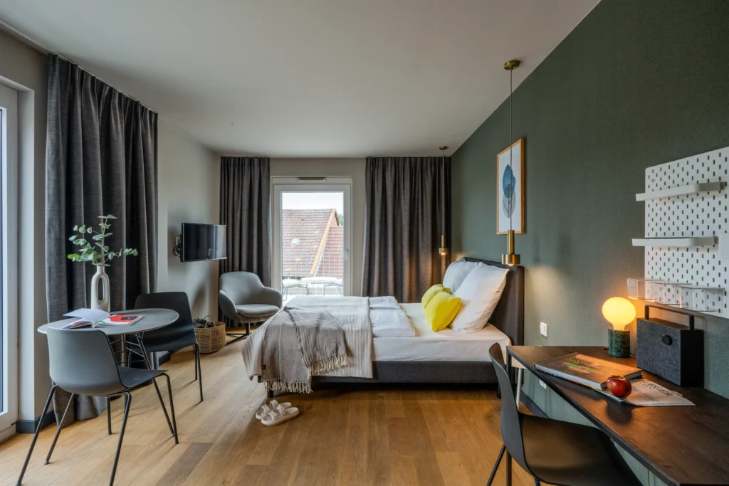 Double bed, armchair, round dining table and desk, window door to the balcony, ipartment Wolfsburg
