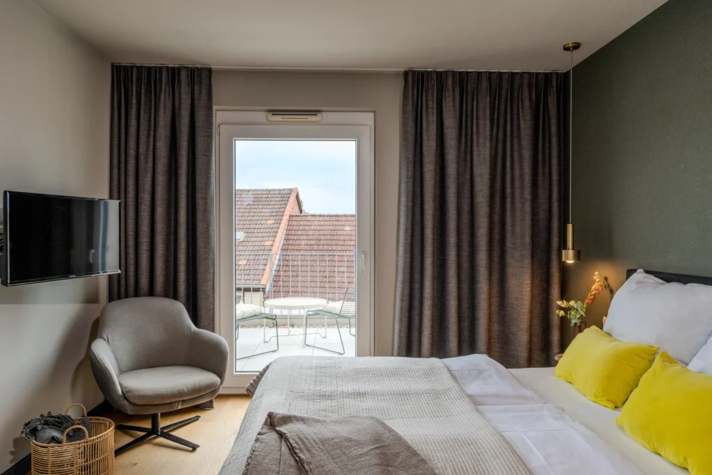 Double bed and armchair in front of the window door with a view of the balcony, ipartment Wolfsburg