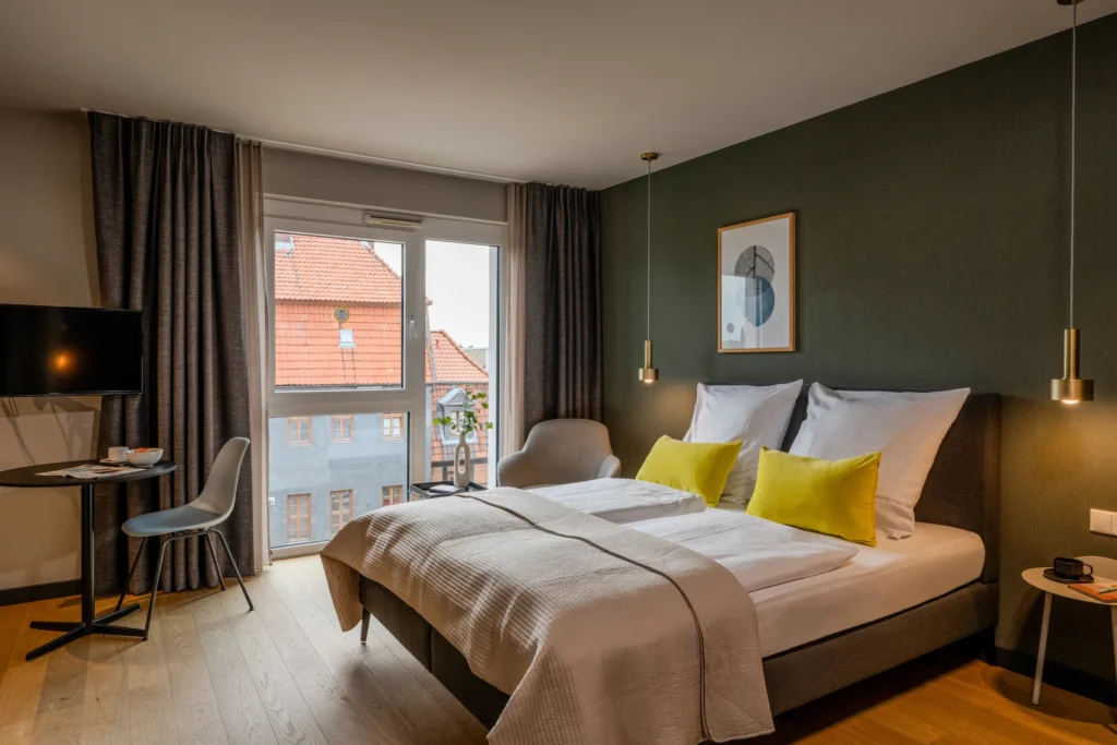 Double bed in front of a green wall, desk and armchair by the window with a view of half-timbered roofs, ipartment Wolfsburg