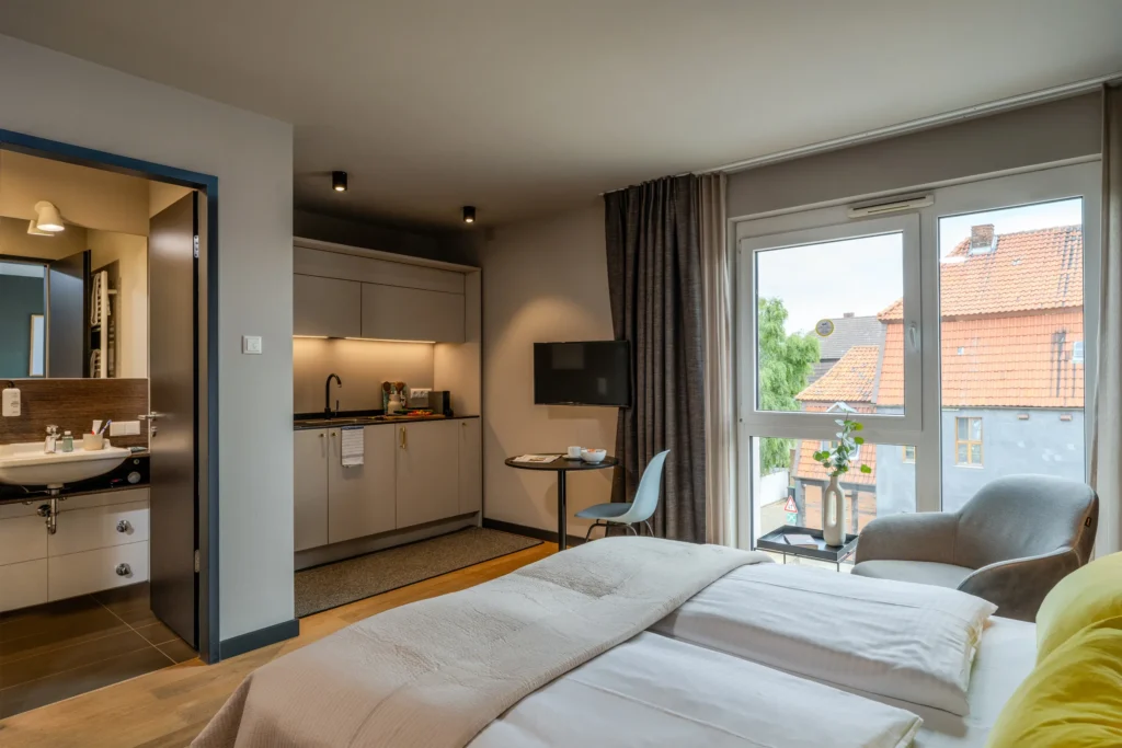 View from the bed over the kitchen area and seating area to the window door; left is the open bathroom door, ipartment Wolfsburg