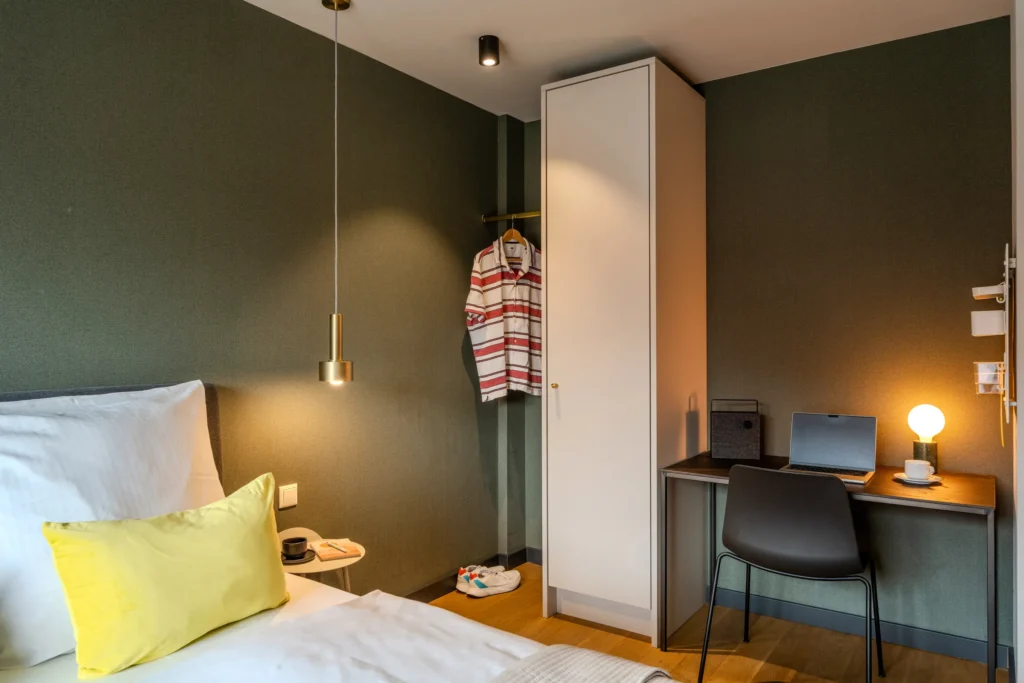 Bed, open wardrobe and desk with laptop in front of green wall, ipartment Wolfsburg