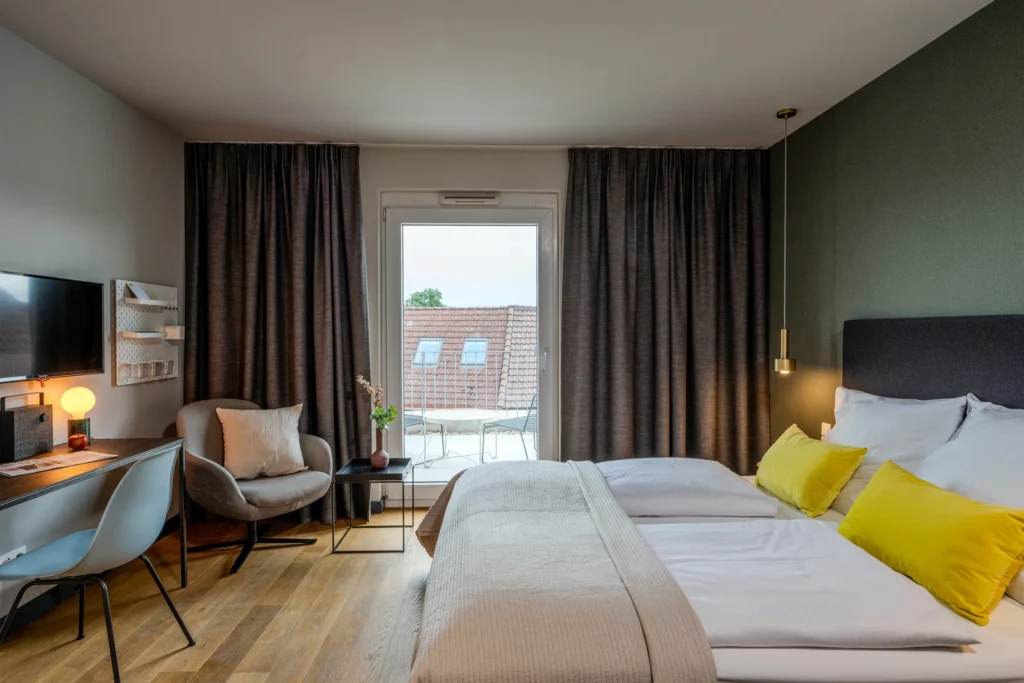 Double bed with a view through the window onto the balcony, armchair in front of it, ipartment Wolfsburg
