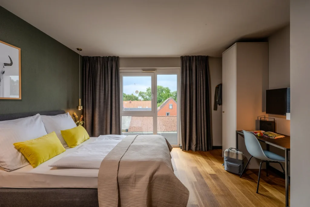 Double bed with yellow pillows, desk and TV, view of half-timbered roofs, ipartment Wolfsburg