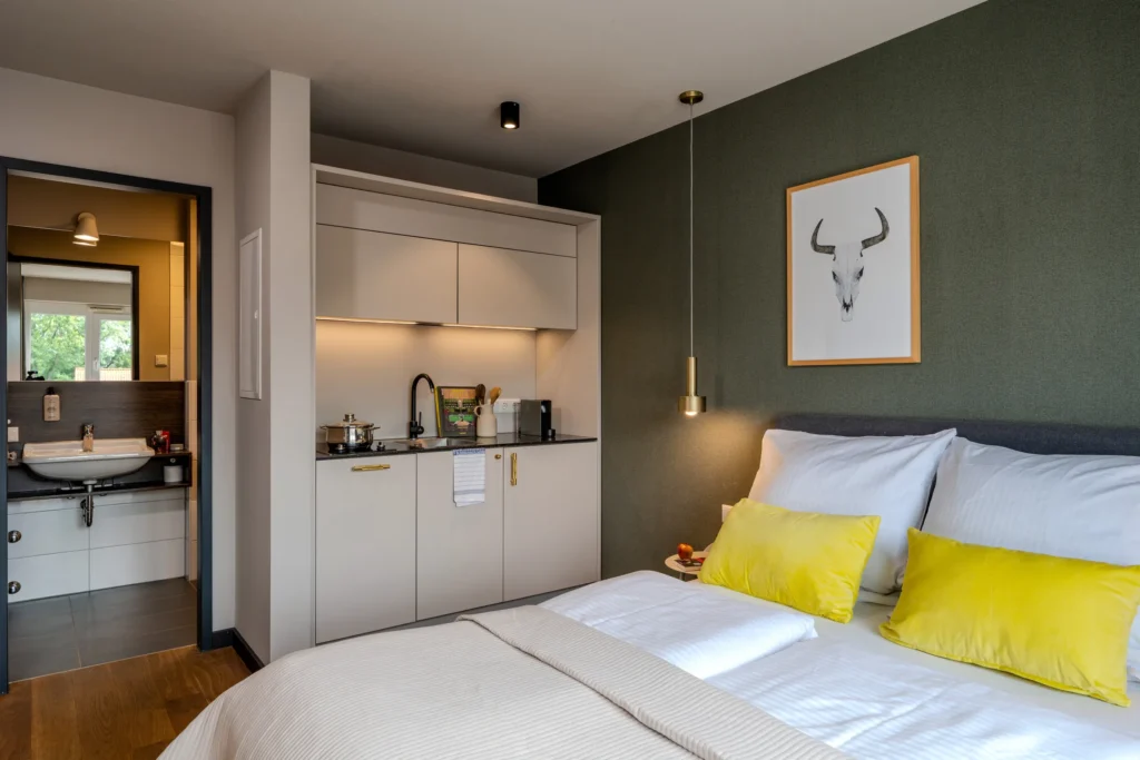 Double bed in front of green wall, kitchenette and open door to the bathroom, ipartment Wolfsburg