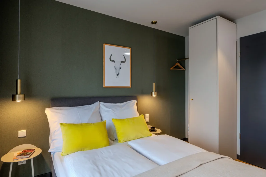 Double bed with yellow pillows in front of a green wall, wardrobe next to it, ipartment Wolfsburg