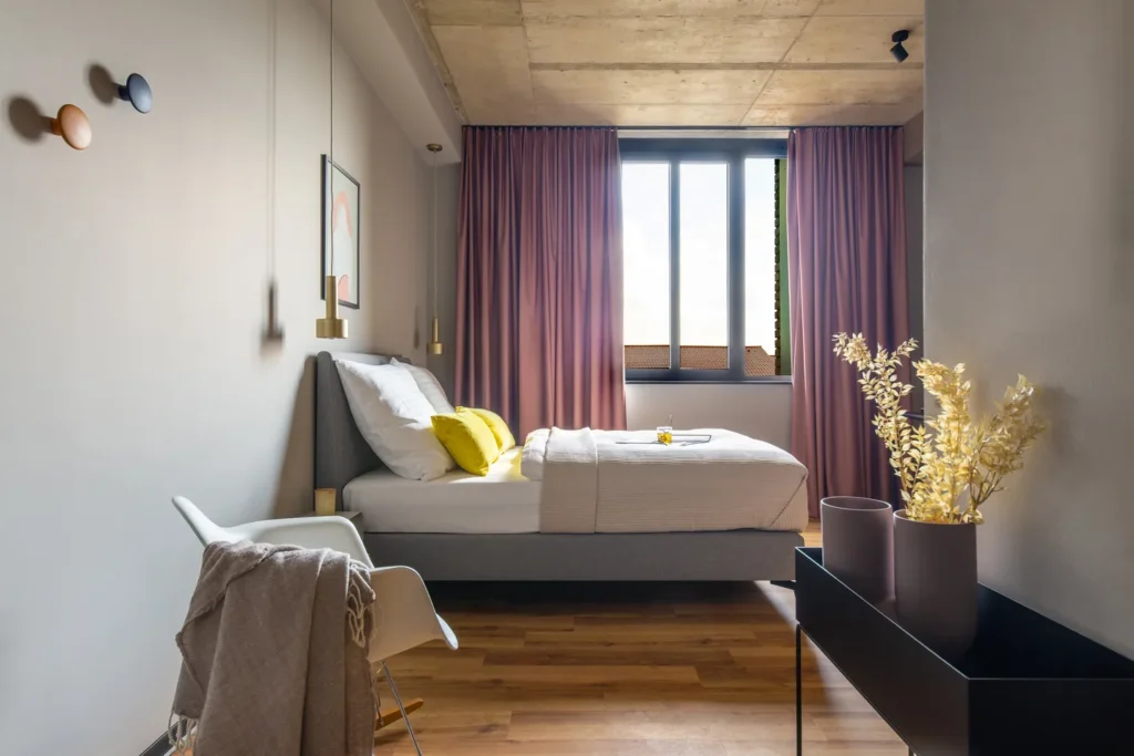 Schlafzimmer mit Doppelbett, rosa Vorhängen und Dekozweigen auf einem schwarzen Sideboard