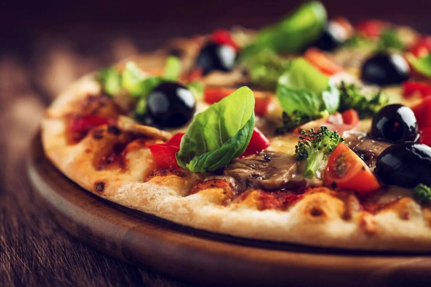 Eine Pizza mit Basilikum, schwarzen Oliven, Tomaten und Pilzen auf einem Holzbrett.